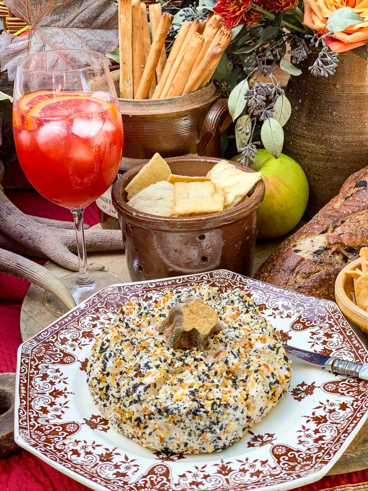 A white and burgandy vintage French plate with pumpkin shaped cheese ball on top, and drinks and snack in the background.