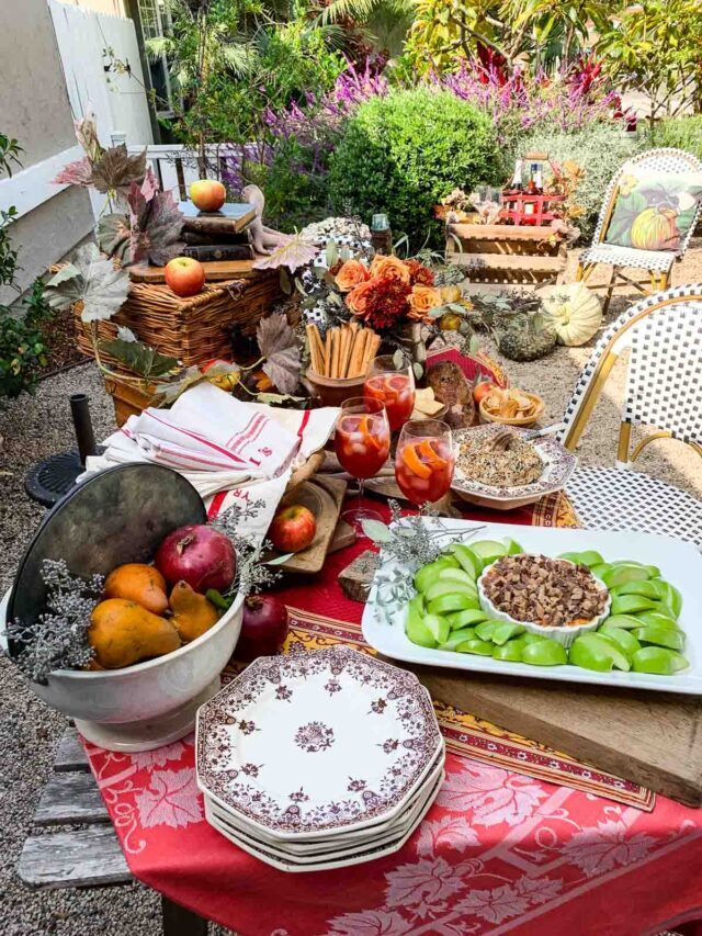 A gorgeous Fall table with French printed red linens and bright colored appetizers featuring and caramel apple dip, pumpkin shaped everything bagel cheese ball, and aperol cocktails at a Fall appetizer outdoor party.