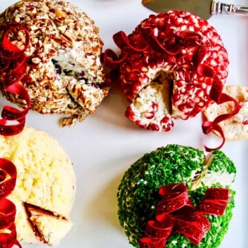 A white square platter with four colorful round cheese balls decorated like Christmas tree ornaments with crackers and spreader knife.