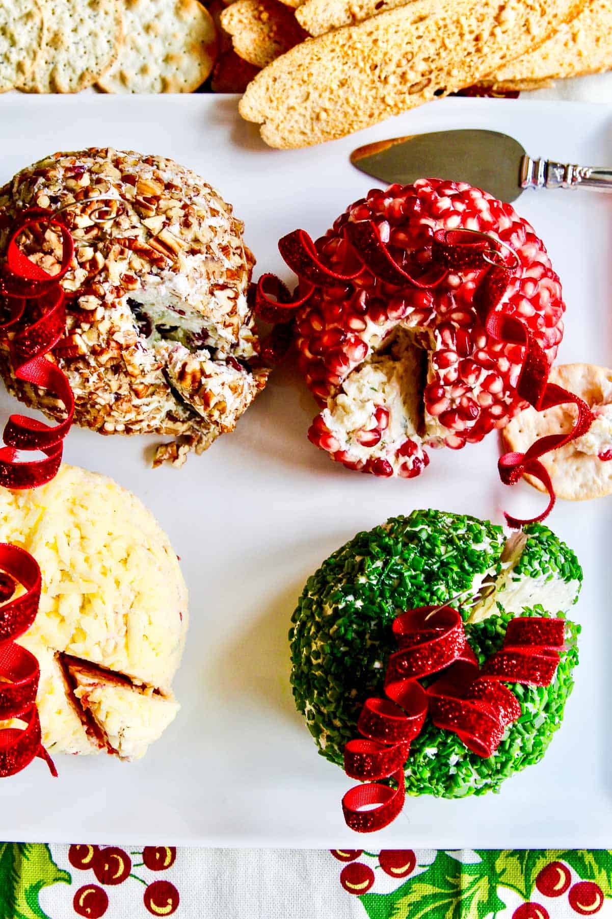 A square white platter with 4 colorful holiday cheese balls shaped like ornaments with crackers and a serving knife on the plate. 