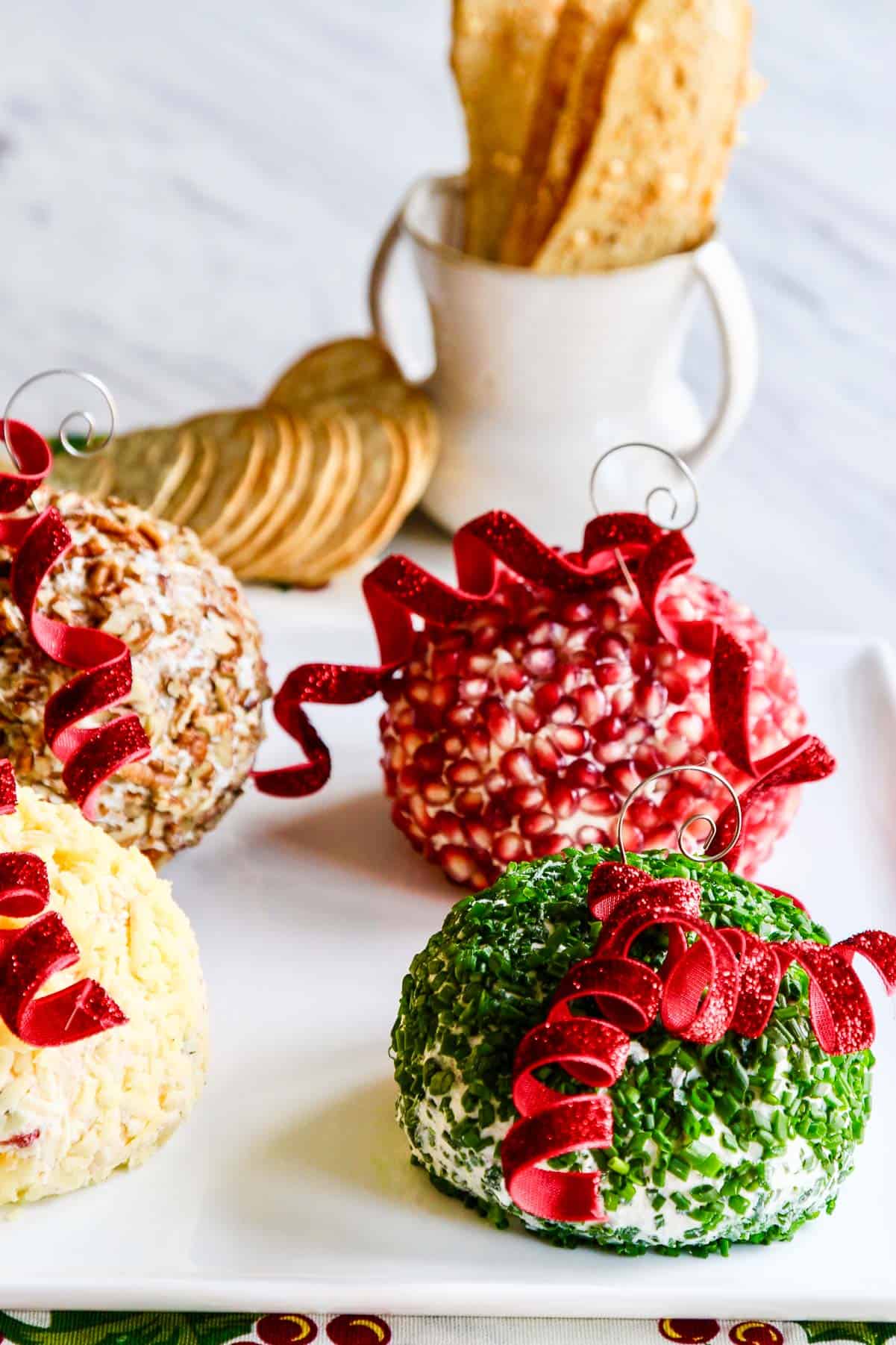 Four bright colored cheese balls decorated to look like tree ornaments with crackers on a white platter.