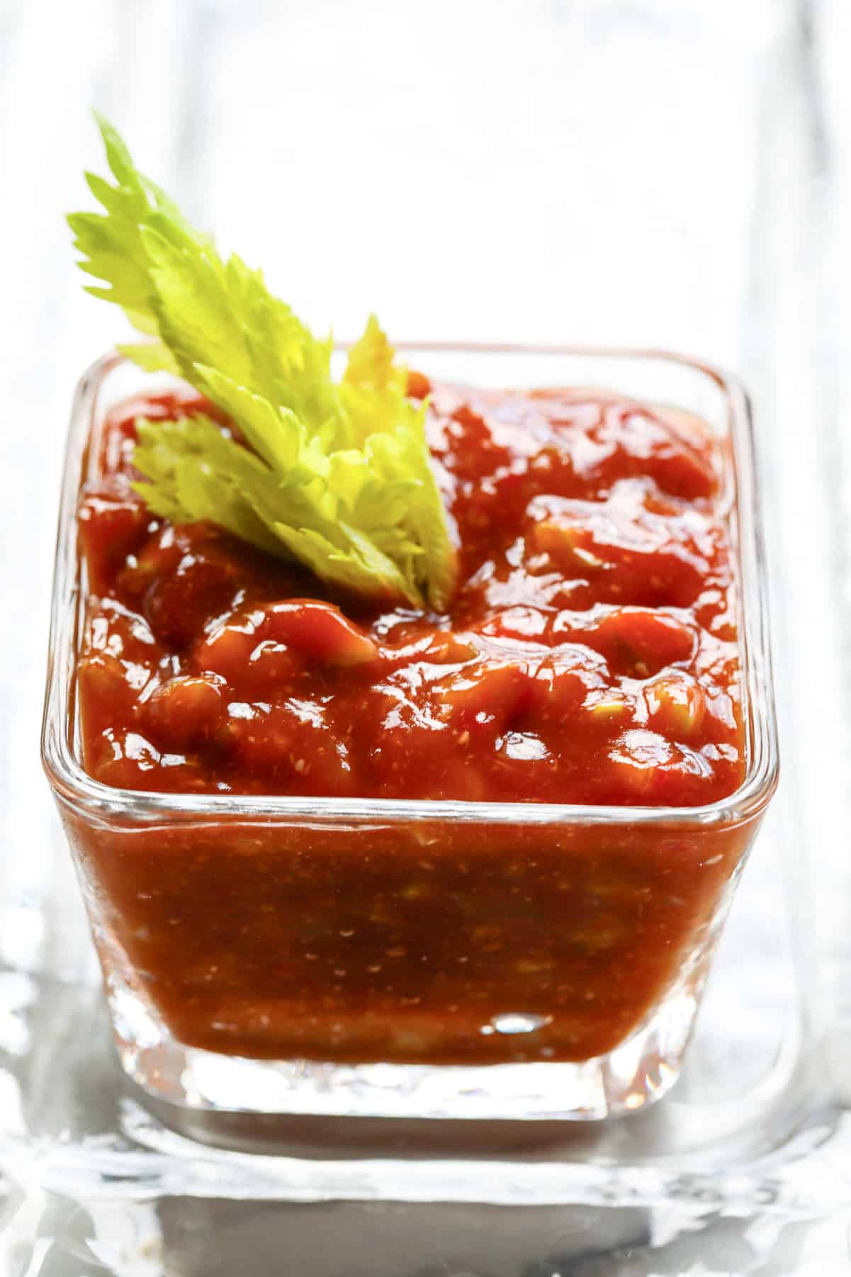 A small square glass bowl filled with homemade cocktail sauce and a celery top garnish to serve with shrimp cocktail.