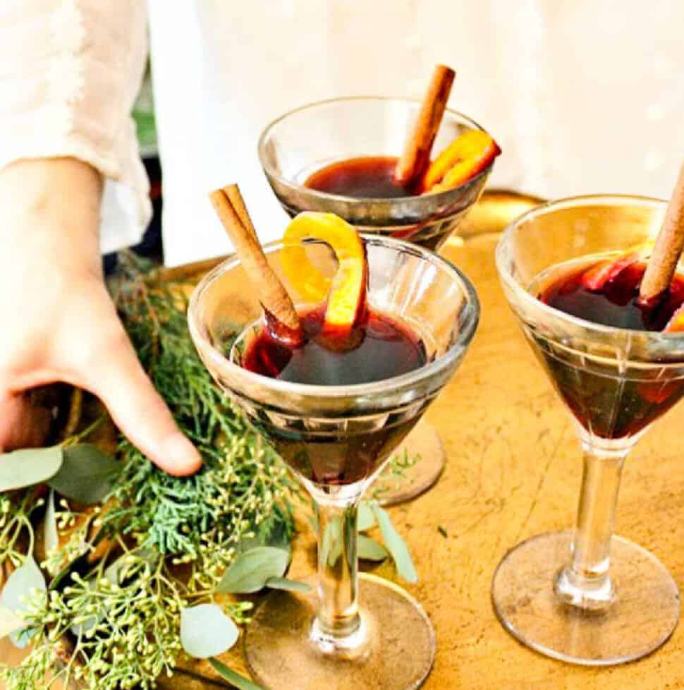 A lady holding a tray of mulled wine garnished with cinnamon sticks and orange peel.