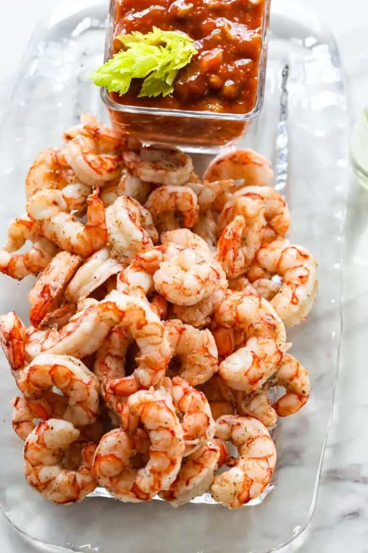 A clear glass platter full of cooked shrimp for shrimp cocktail with cocktail sauce nearby.