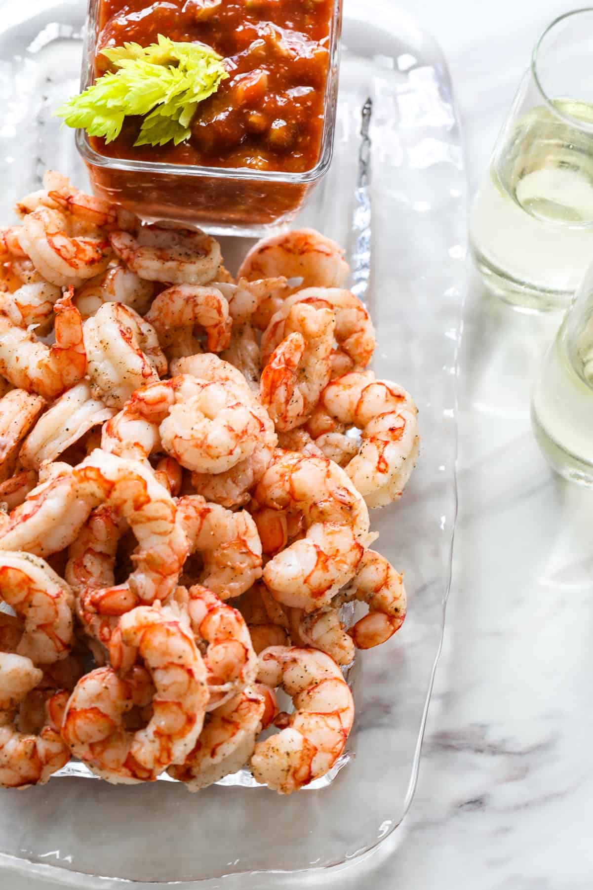 A clear glass platter full of cooked shrimp for shrimp cocktail with cocktail sauce nearby.