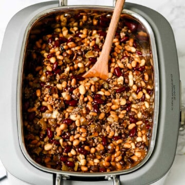 A large slow cooker filled with canned beans, chopped onions, and hamburger to make Cowboy Baked Beans a recipe for a large party.