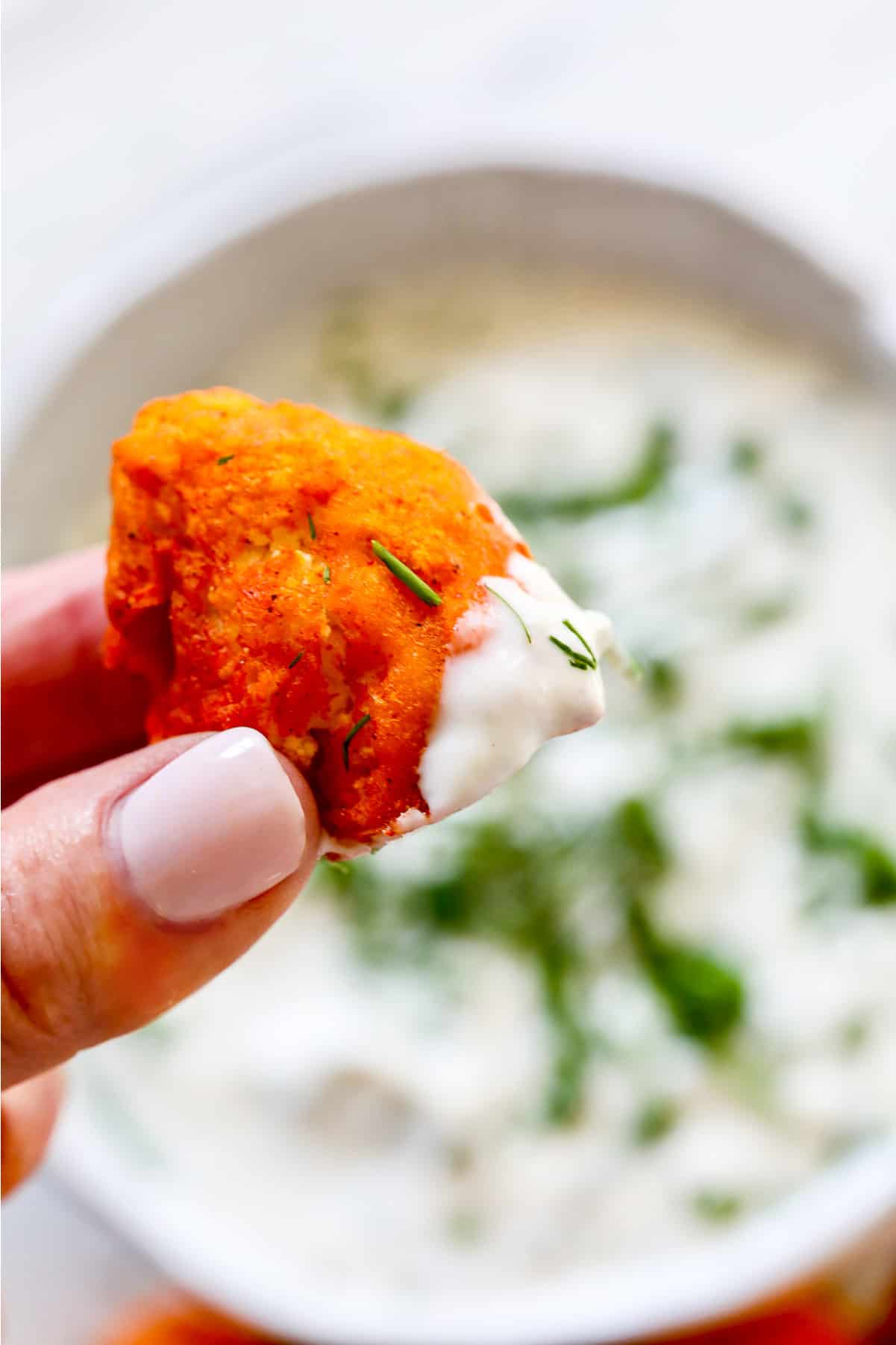 A lady holding a piece of buffalo cauliflower "wing" over a bowl of ranch dip in the background.