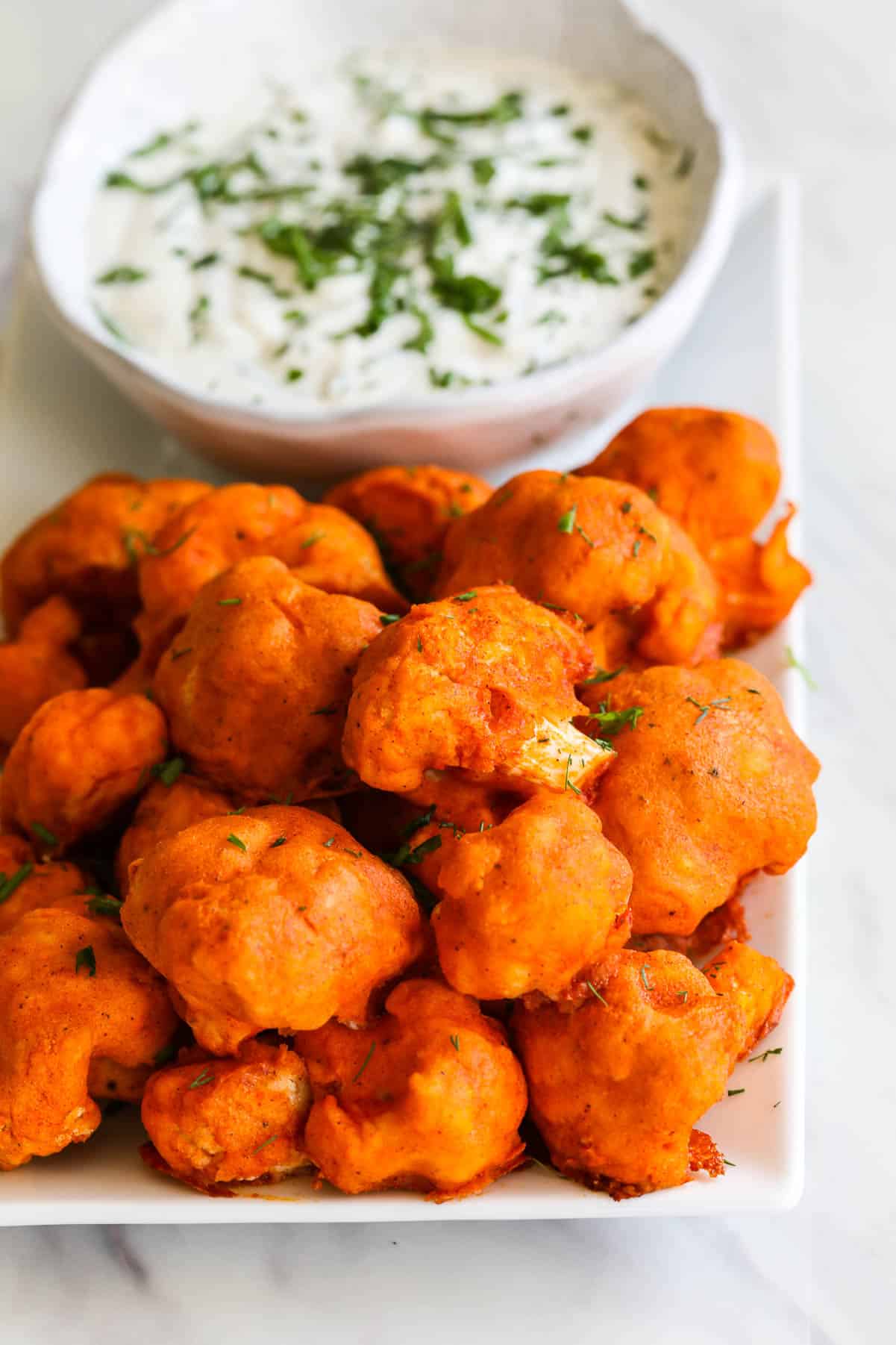 A pile of baked Buffalo Cauliflower on a plate with ranch dip sprinkled with fresh herbs.