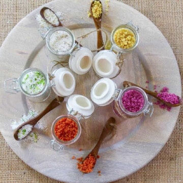 A wood plate with colorful homemade salts and seasoning spice blends in small glass jars with spoons.