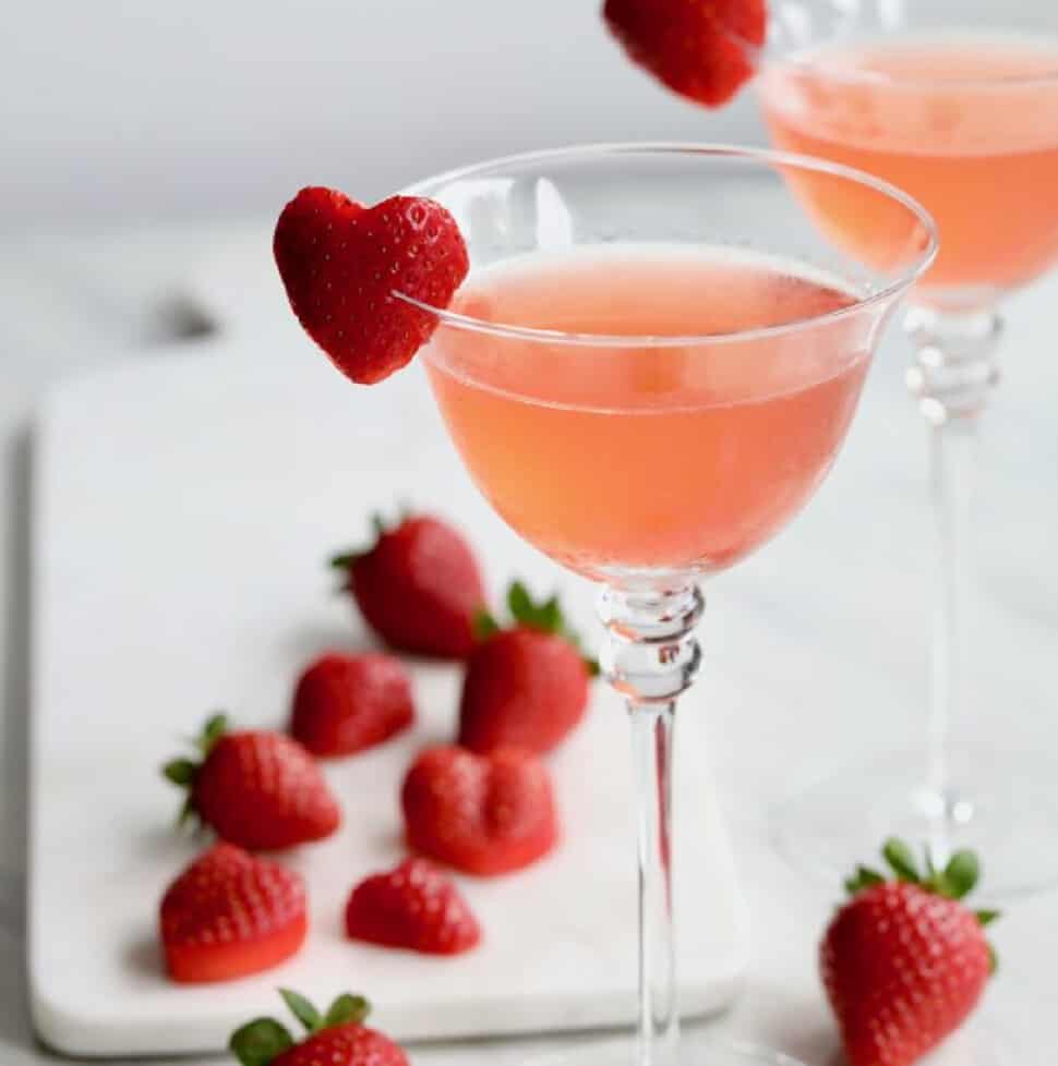 A pink cocktail with a sliced strawberry in the shape of a heart on the edge of the glass as a garnish.