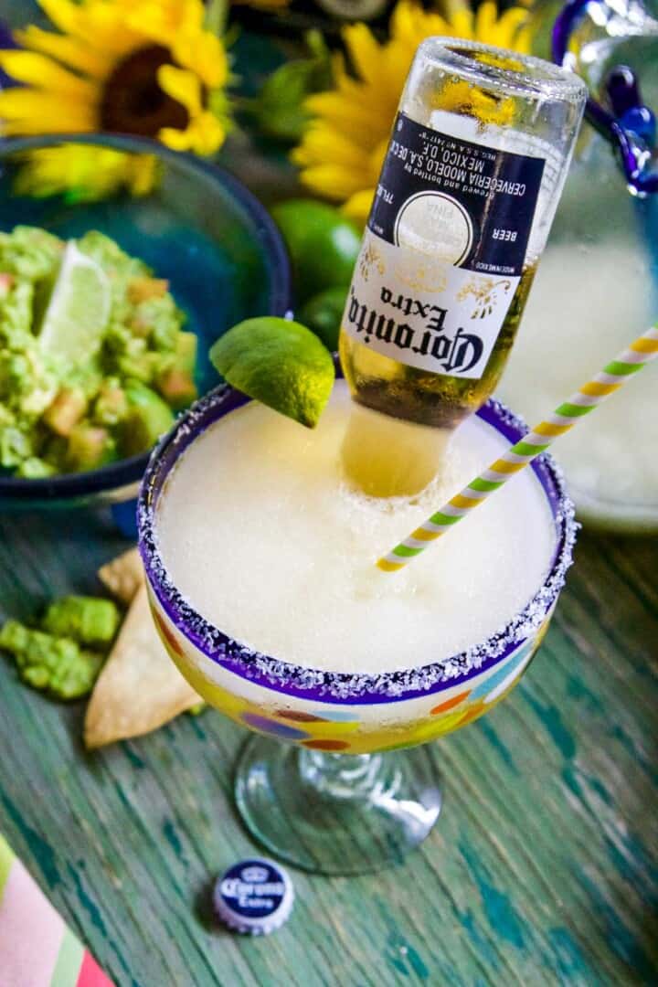 A large blue rimmed margarita glass with straw, lime, and Coronita beer poured inside the glass.