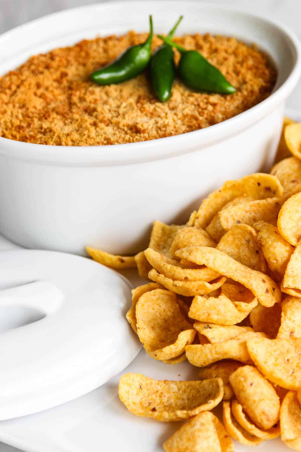 A white casserole dish with jalapeno popper dip garnished with 3 green jalapenos and large corn chips.