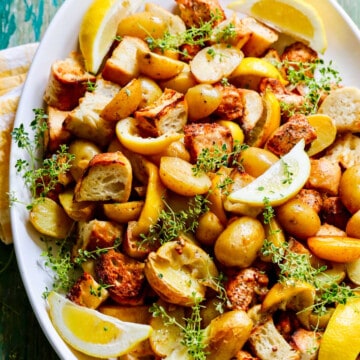 A white platter filled with sheet pan chicken dinner including lemons, potatoes, and croutons.