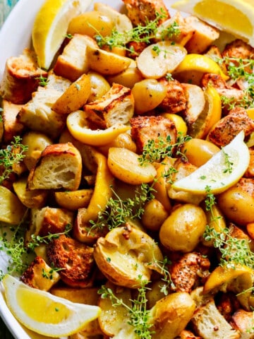 A white platter filled with sheet pan chicken dinner including lemons, potatoes, and croutons.