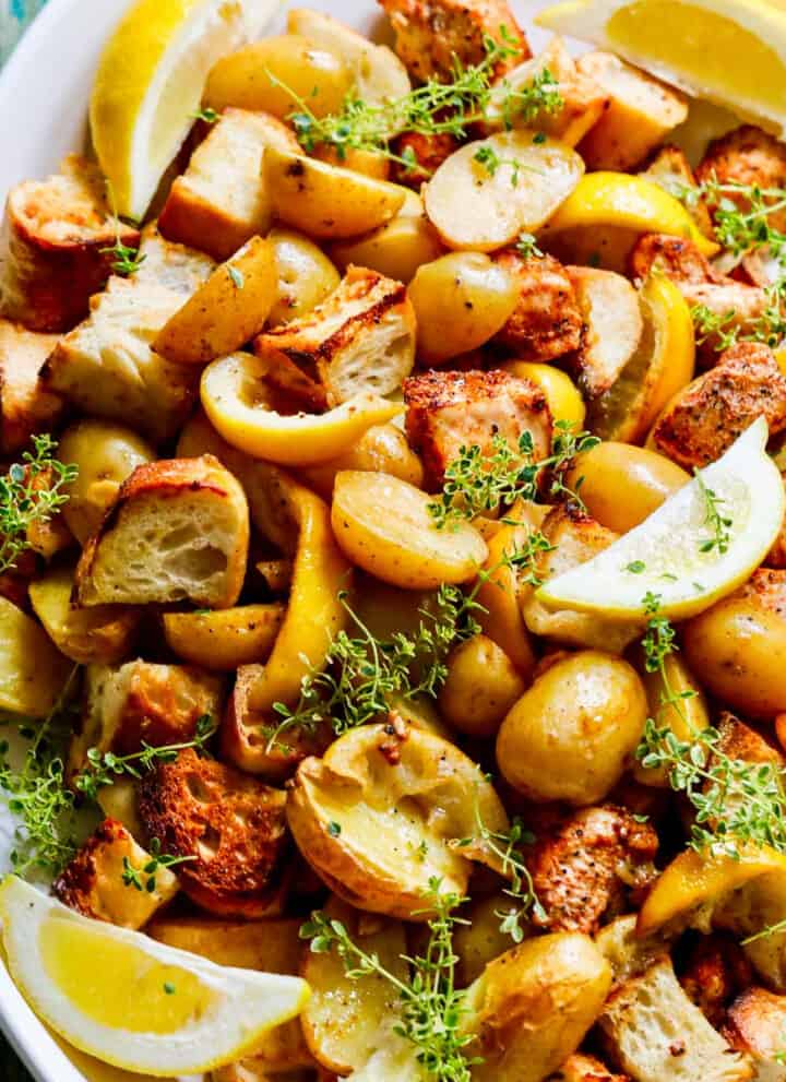 A white platter filled with sheet pan chicken dinner including lemons, potatoes, and croutons.