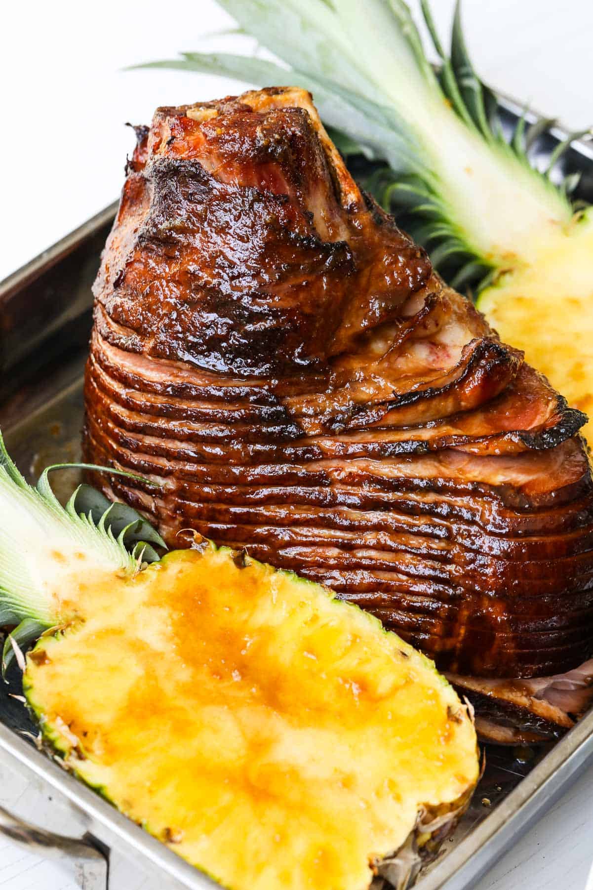A bone in glazed ham with a whole pineapple cut in half in a roasting pan.