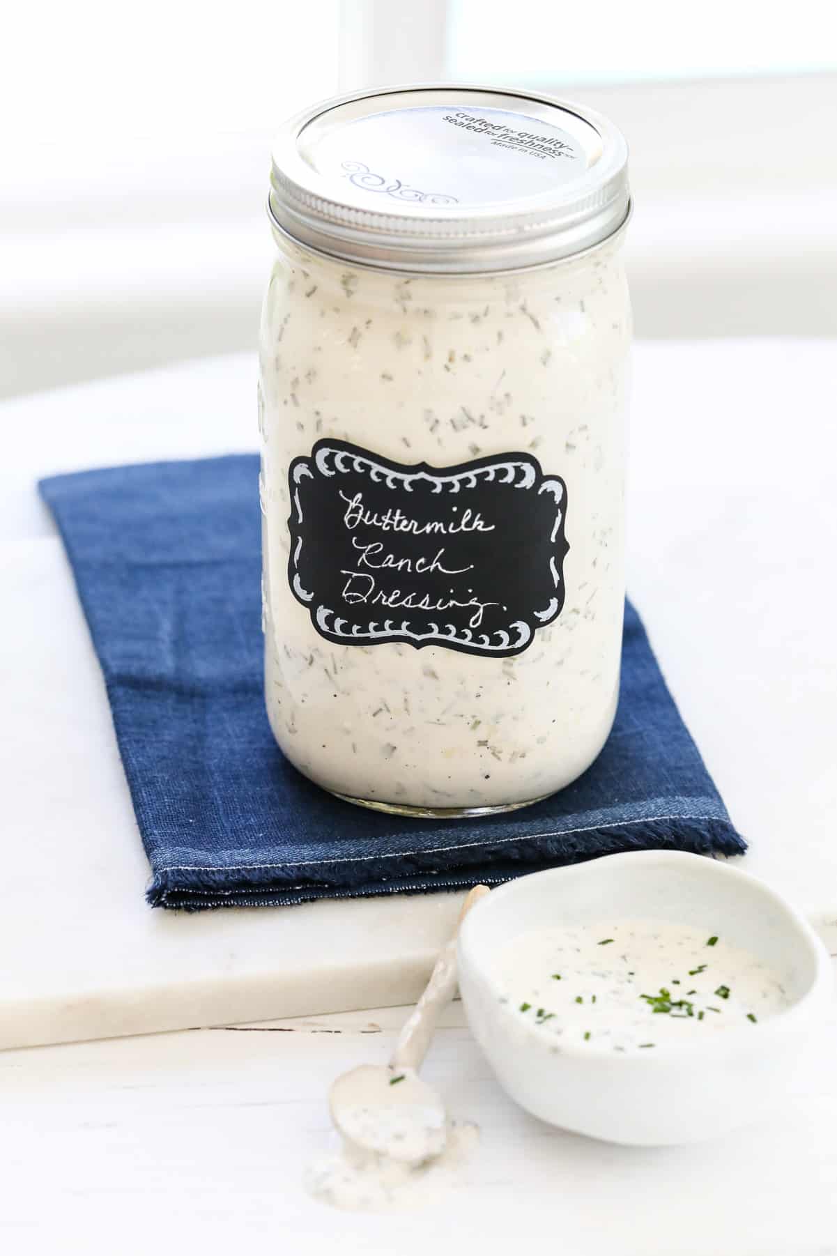 A large Mason jar filled with Ranch Dressing made at home on a blue napkin with a small bowl and spoon.
