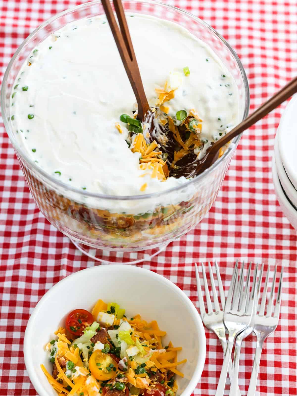 A glass trifle dish filled with colorful 7 Layer Salad being served with wooden tongs.