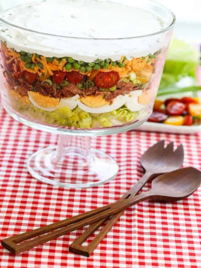 A large glass trifle bowl filled with a 7 Layer Salad with wood tong on the table and a red and white check cloth.