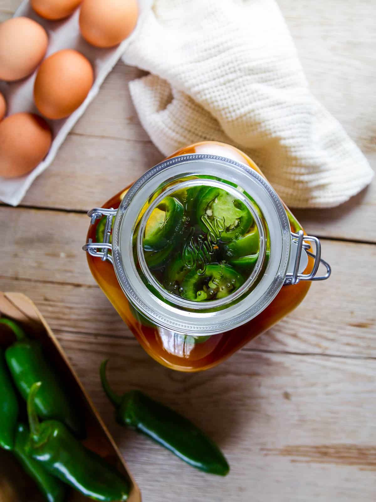 Looking down on a jar of pickled eggs with a carton of eggs and basket of jalapenos.