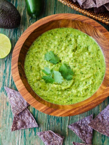 A wood bowl with green avocado salsa taco sauce and blue corn tortilla chips on the table.