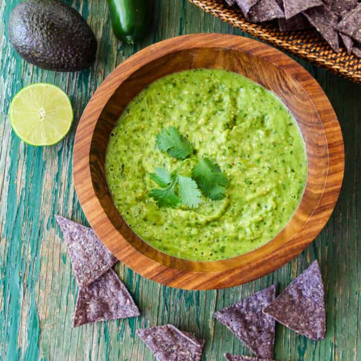 A wood bowl with green avocado salsa taco sauce and blue corn tortilla chips on the table.