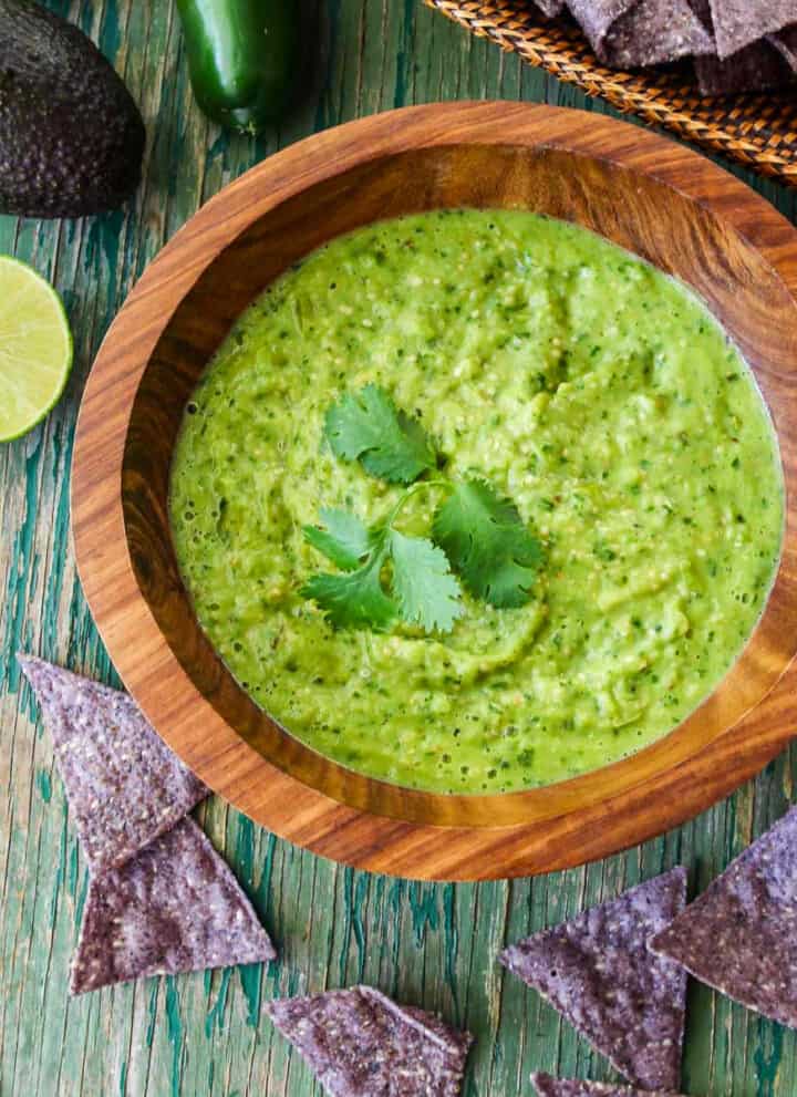 A wood bowl with green avocado salsa taco sauce and blue corn tortilla chips on the table.