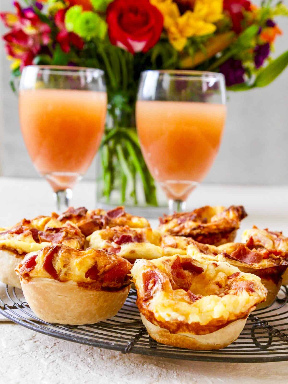 Two glasses of grapefruit juice with mini quiches and flowers in the background at brunch.