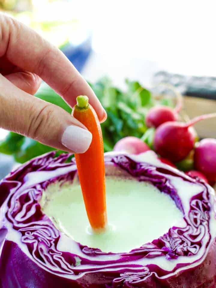 A purple cabbage filled with dip and a lady dipping a small orange carrot into it.