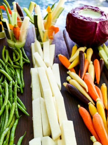 A cutting board with crudites and dip with bright colored vegetables cut into strips and sticks.