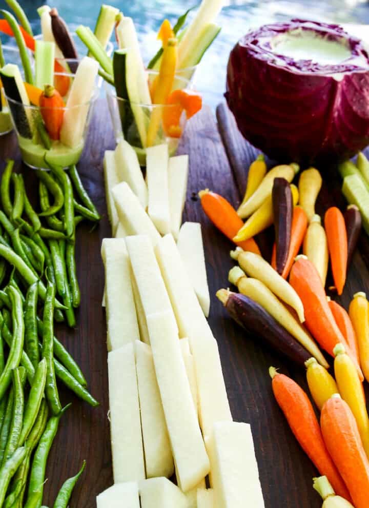 A cutting board with crudites and dip with bright colored vegetables cut into strips and sticks.