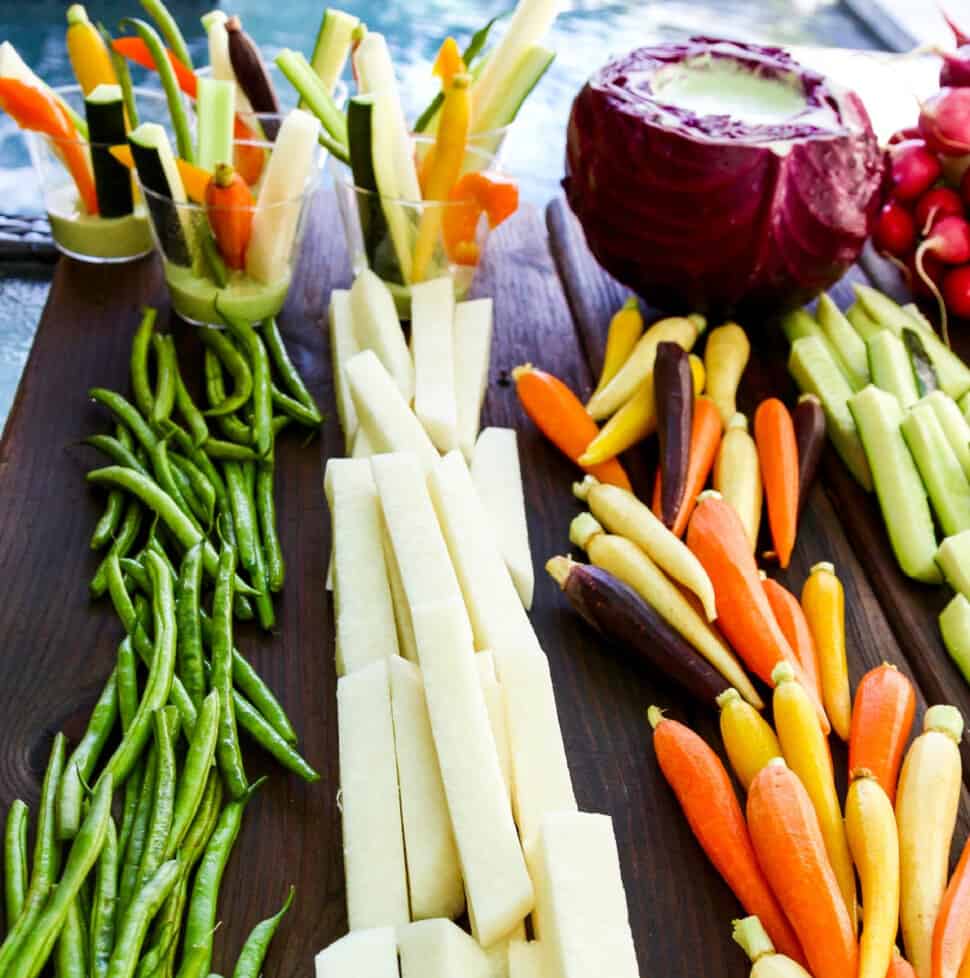 A cutting board with crudites and dip with bright colored vegetables cut into strips and sticks.