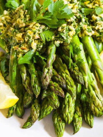 A white plate filled with cooked asparagus topped with gremolata garnished with lemon wedges.