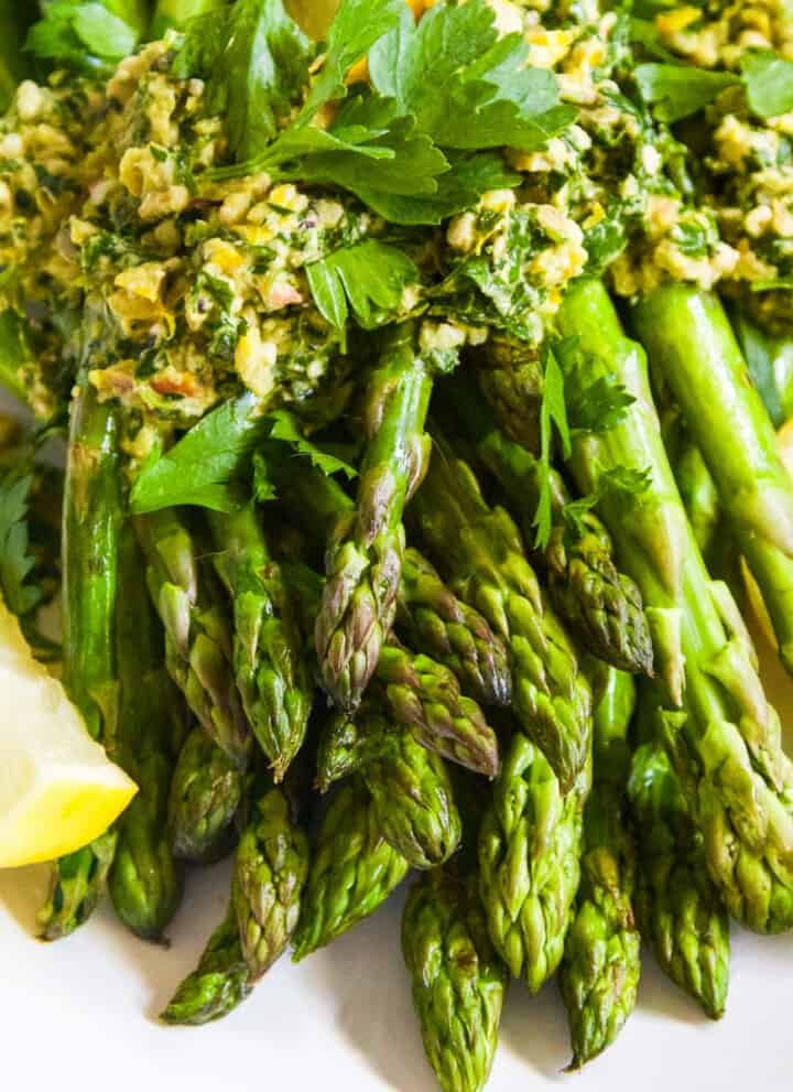 A white plate filled with cooked asparagus topped with gremolata garnished with lemon wedges.