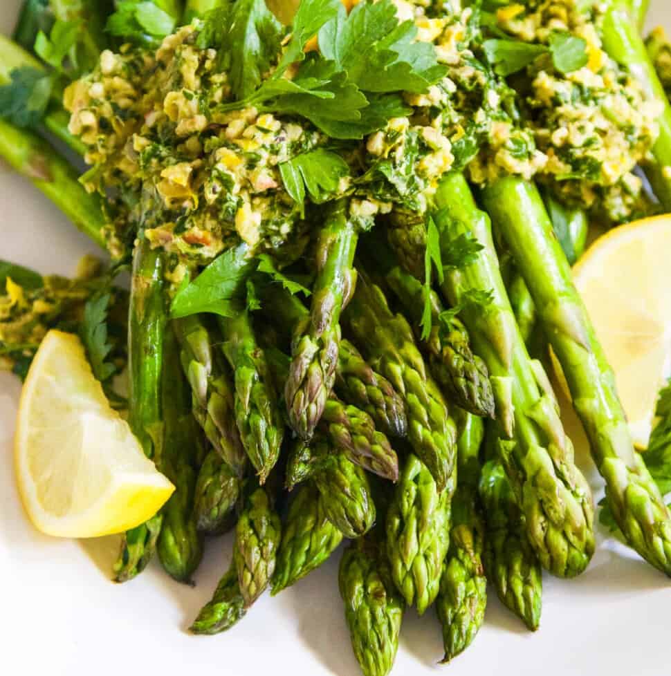 A white plate filled with cooked asparagus topped with gremolata garnished with lemon wedges.