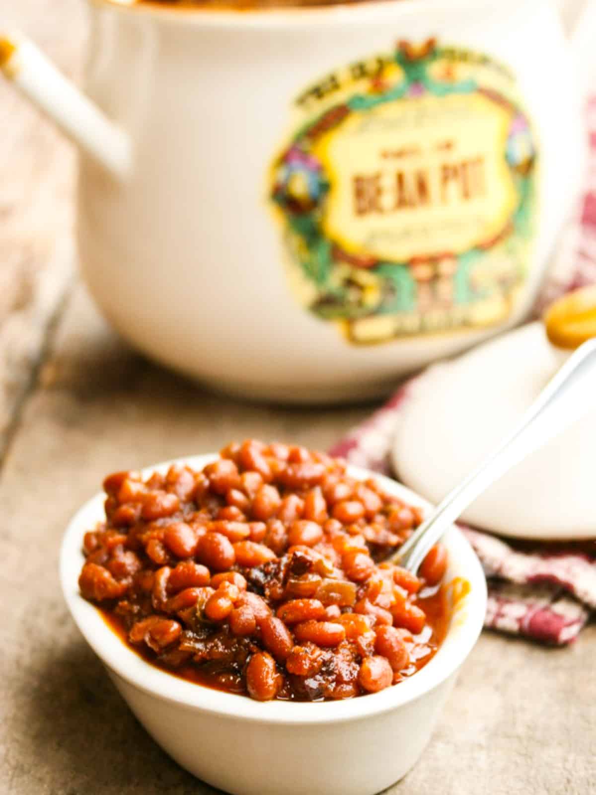 A small white bowl filled with baked beans with a bean pot in the background.