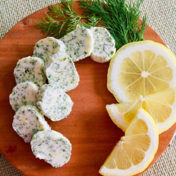 Small brown round cutting board with slices of lemon dill butter garnished with fresh lemon and dill.