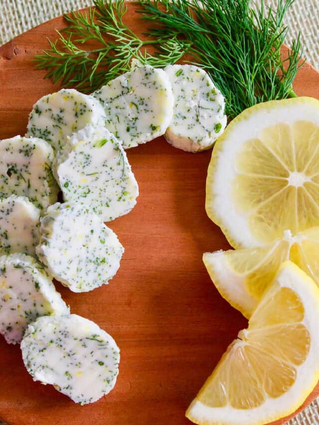 Small brown round cutting board with slices of lemon dill butter garnished with fresh lemon and dill.