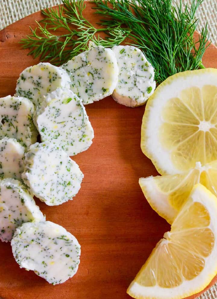 Small brown round cutting board with slices of lemon dill butter garnished with fresh lemon and dill.