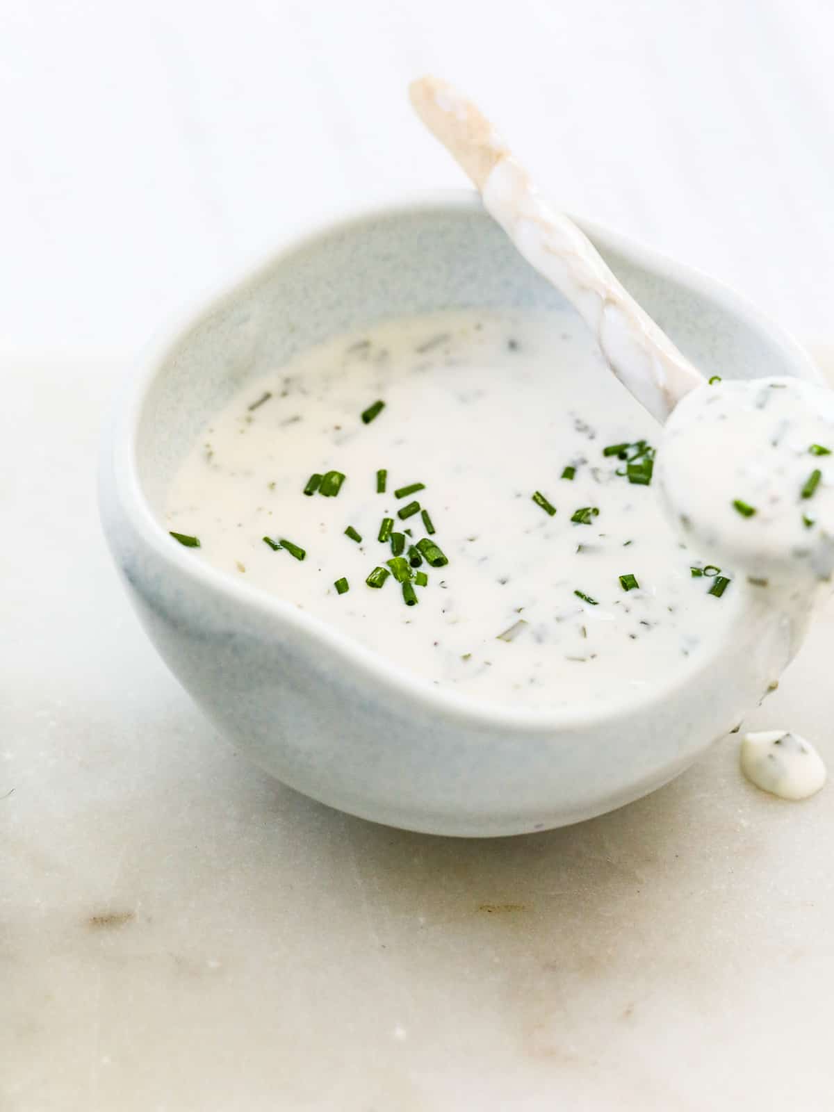A small white bowl filled with ranch dressing and a tiny spoon to use on a salad.