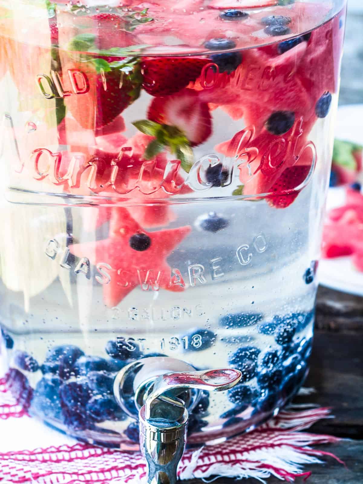 A large glass dispenser filled with infused water garnished with blueberries, watermelon stars, and strawberries.