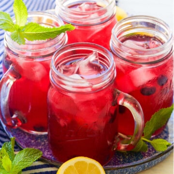 Mason jar handled glasses with blueberry lemonade garnished with blueberries, mint, and lemons halves.