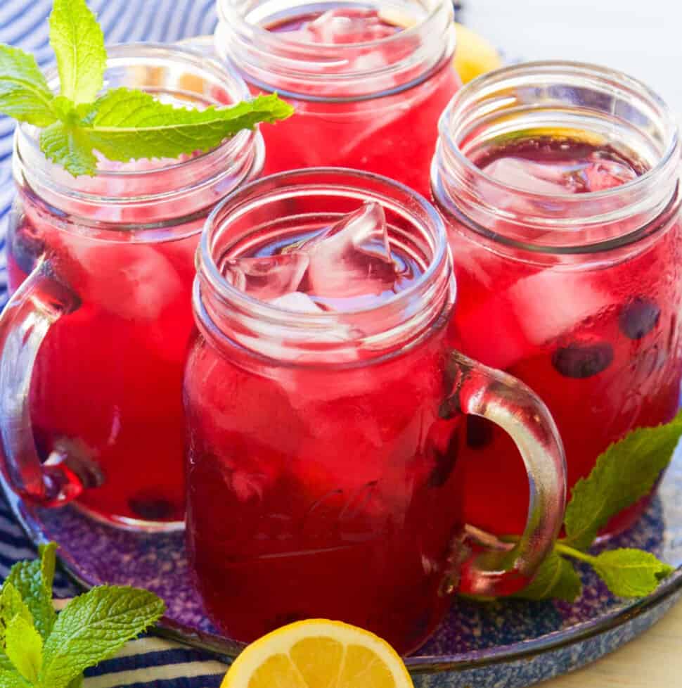 Mason jar handled glasses with blueberry lemonade garnished with blueberries, mint, and lemons halves.