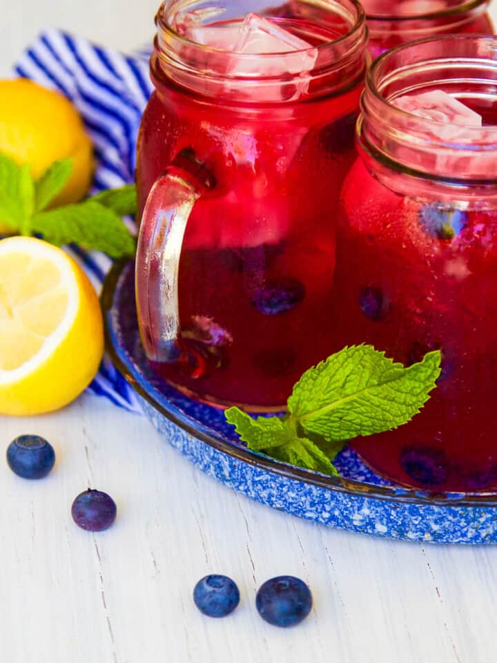 Mason jar handled glasses with blueberry lemonade garnished with blueberries, mint, and lemons halves.