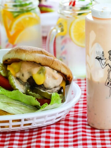 A messy grilled burger topped with lettuce, tomato, cheese, pickles and burger sauce in a basket with a bottle of burger sauce.