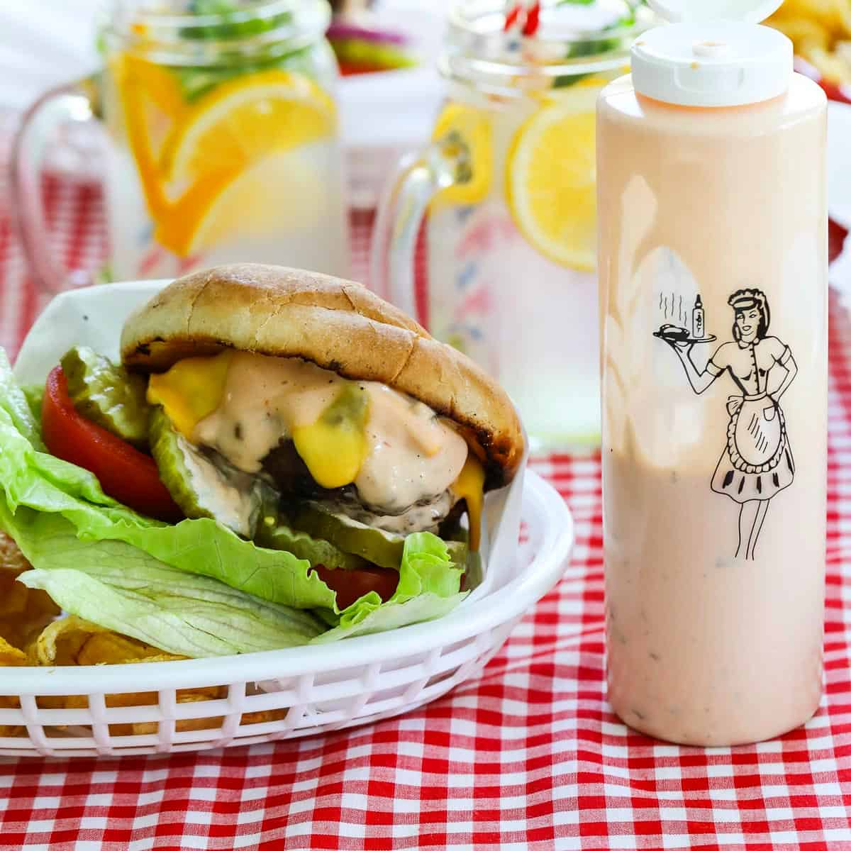 A messy grilled burger topped with lettuce, tomato, cheese, pickles and burger sauce in a basket with a bottle of burger sauce.