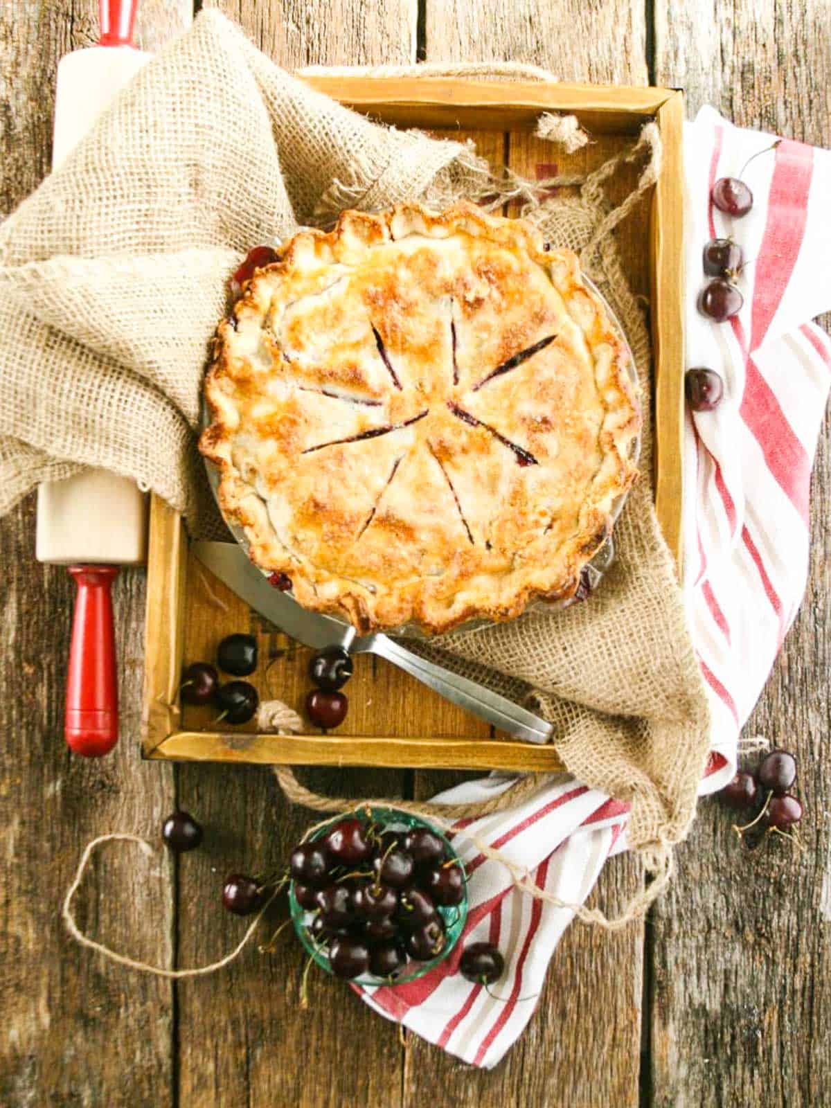 A large cherry pie with rolling pin and bowl of cherries ready to cut a slice.