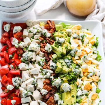 A white square plate filled with colorful Cobb Salad made with chicken, eggs, avocados, tomatoes, and bacon.