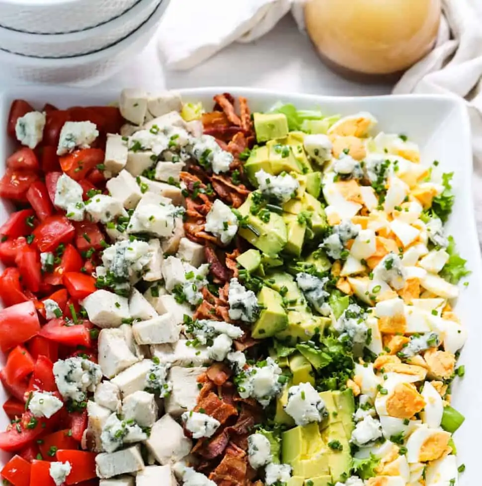 A white square plate filled with colorful Cobb Salad made with chicken, eggs, avocados, tomatoes, and bacon.