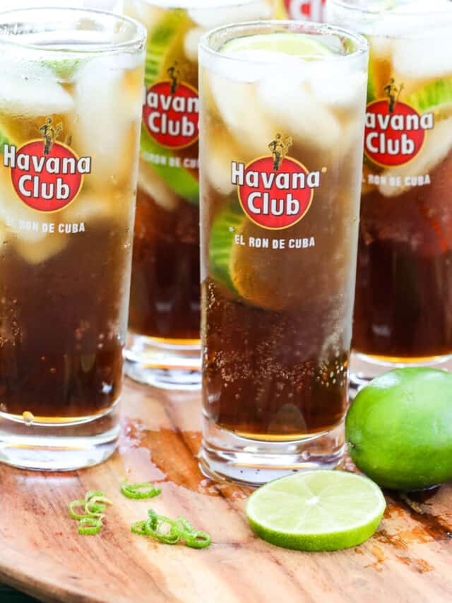 Four tall Havana Club glasses with Cuba Libre cocktails garnished with lime on a wood tray.